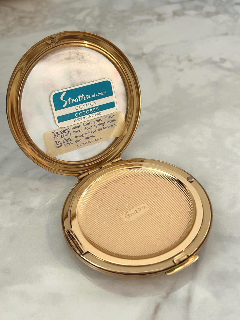 May include: A round gold-tone metal powder compact with an open lid. The interior features a mirror with a label reading Stratton of London COSMOS OCTOBER MADE IN ENGLAND and instructions for use. The base has a textured gold-tone metal surface.