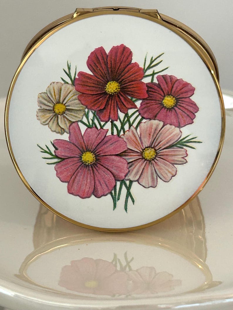 May include: A round gold-tone metal compact mirror featuring a white lid decorated with a colorful illustration of five cosmos flowers in shades of deep red, magenta, light pink, and white, each with a yellow center and green foliage.