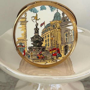 May include: A round gold-tone metal compact mirror featuring a colorful illustration of Piccadilly Circus in London. The scene depicts the Eros statue, a red double-decker bus, and the text Piccadilly + Eros printed in black at the bottom.