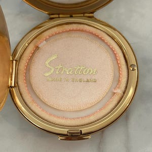 May include: A gold-tone metal powder compact with a hinged lid and a circular mirror. The interior features a pale pink powder puff with the gold text Stratton MADE IN ENGLAND. The mirror lid includes instructions for opening and closing the compact.