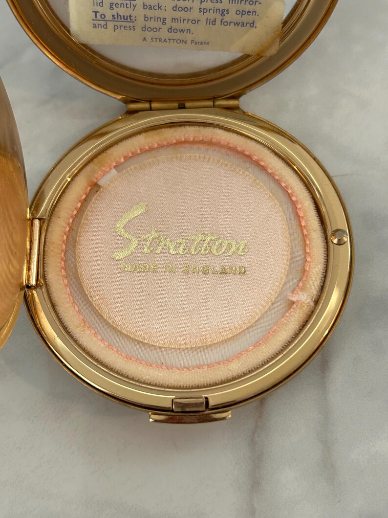 May include: A gold-tone metal powder compact with a hinged lid and a circular mirror. The interior features a pale pink powder puff with the gold text Stratton MADE IN ENGLAND. The mirror lid includes instructions for opening and closing the compact.