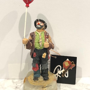 May include: A ceramic clown figurine holding a red balloon with blue polka dots. The clown wears a patched outfit, a hat, and has a painted face. The figurine stands on a stone base with gold glitter.