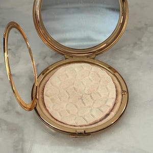 Peut inclure: Un bo&icirc;tier compact rond en m&eacute;tal dor&eacute; dot&eacute; d'un couvercle &agrave; charni&egrave;re avec un miroir circulaire int&eacute;gr&eacute;. &Agrave; l'int&eacute;rieur, une poudre press&eacute;e circulaire pr&eacute;sente un motif g&eacute;om&eacute;trique en nid d'abeille en relief sur sa surface beige clair.