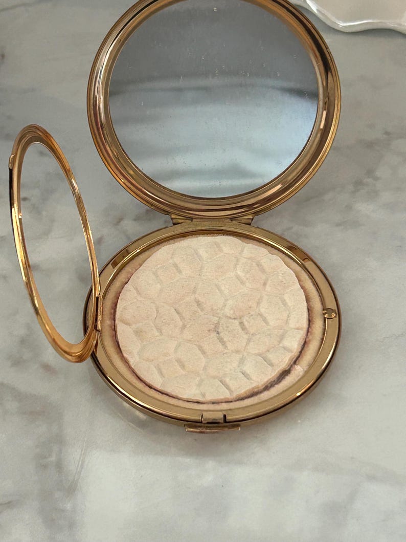 Peut inclure: Un bo&icirc;tier compact rond en m&eacute;tal dor&eacute; dot&eacute; d'un couvercle &agrave; charni&egrave;re avec un miroir circulaire int&eacute;gr&eacute;. &Agrave; l'int&eacute;rieur, une poudre press&eacute;e circulaire pr&eacute;sente un motif g&eacute;om&eacute;trique en nid d'abeille en relief sur sa surface beige clair.