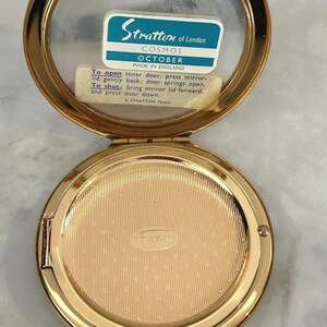 May include: A gold-tone metal Stratton of London Cosmos October compact mirror with an open lid. The interior features a mirror with a blue and white label reading Stratton of London Cosmos October Made in England and instructions for the inner door.