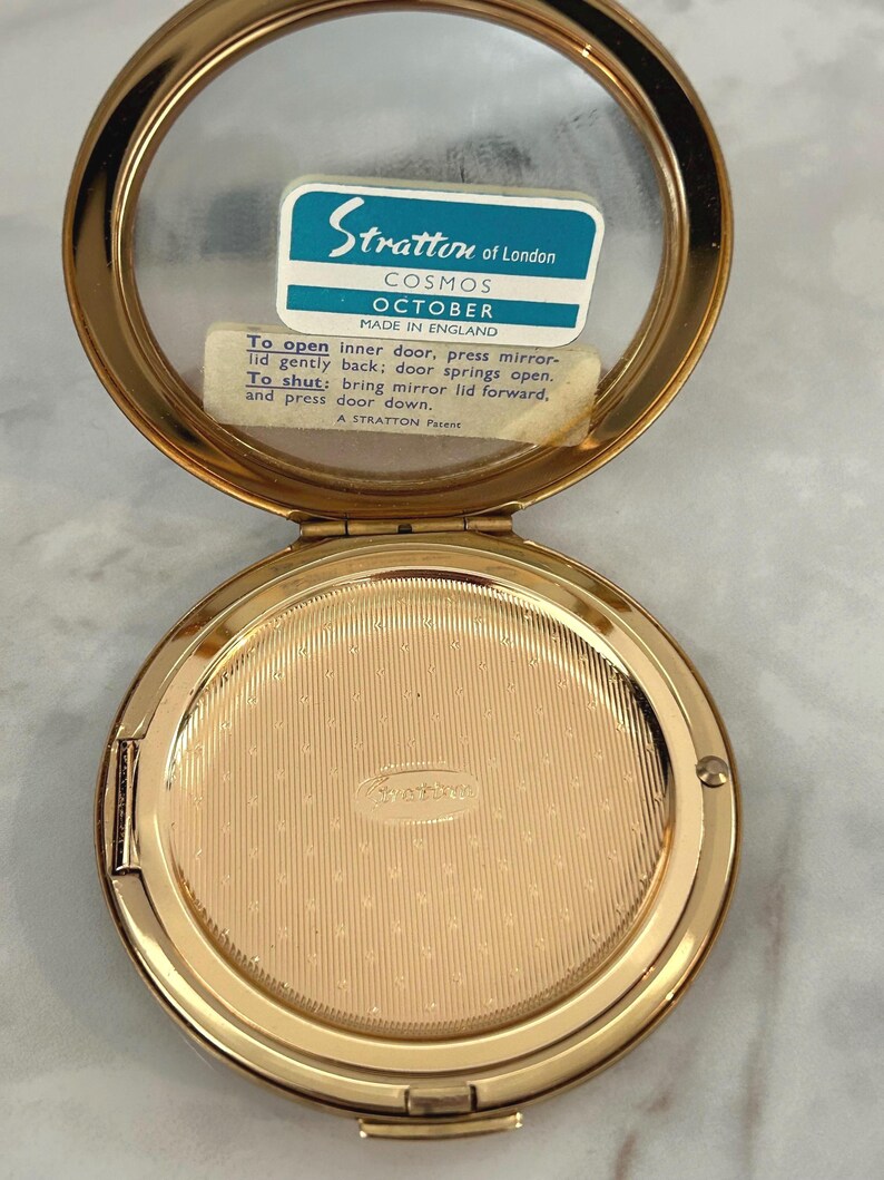 May include: A gold-tone metal Stratton of London Cosmos October compact mirror with an open lid. The interior features a mirror with a blue and white label reading Stratton of London Cosmos October Made in England and instructions for the inner door.