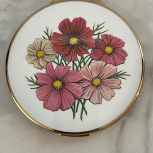 May include: A round compact mirror with a gold-toned metal frame and a white enamel lid. The lid features a central illustration of a bouquet of cosmos flowers in shades of pink, red, and white with yellow centers and green stems.
