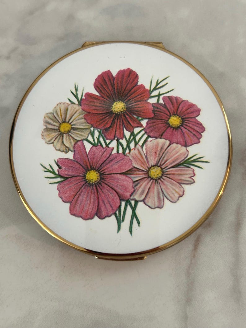 May include: A round compact mirror with a gold-toned metal frame and a white enamel lid. The lid features a central illustration of a bouquet of cosmos flowers in shades of pink, red, and white with yellow centers and green stems.