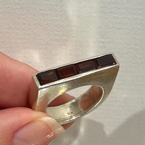 May include: A sterling silver ring featuring a flat, rectangular top bar set with four deep red, rectangular-cut garnet gemstones. The band has a brushed metal finish and is stamped with the number 925 on the interior surface of the ring.