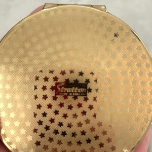 May include: A round gold-tone metal compact mirror featuring a textured surface with a repeating pattern of small stars. The center displays a rectangular black and red label with the text Stratton MADE IN ENGLAND printed in white and gold lettering.