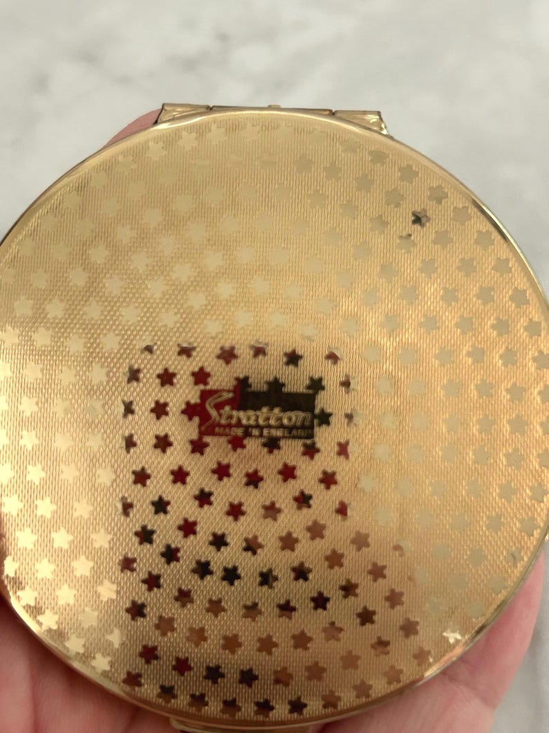 May include: A round gold-tone metal compact mirror featuring a textured surface with a repeating pattern of small stars. The center displays a rectangular black and red label with the text Stratton MADE IN ENGLAND printed in white and gold lettering.