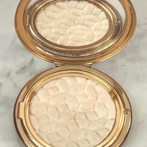 Peut inclure: Un bo&icirc;tier compact rond de couleur dor&eacute;e avec un couvercle &agrave; charni&egrave;re et un miroir int&eacute;gr&eacute;. L'int&eacute;rieur contient une poudre press&eacute;e pr&eacute;sentant un motif g&eacute;om&eacute;trique en relief rappelant un nid d'abeille, de couleur cr&egrave;me p&acirc;le.