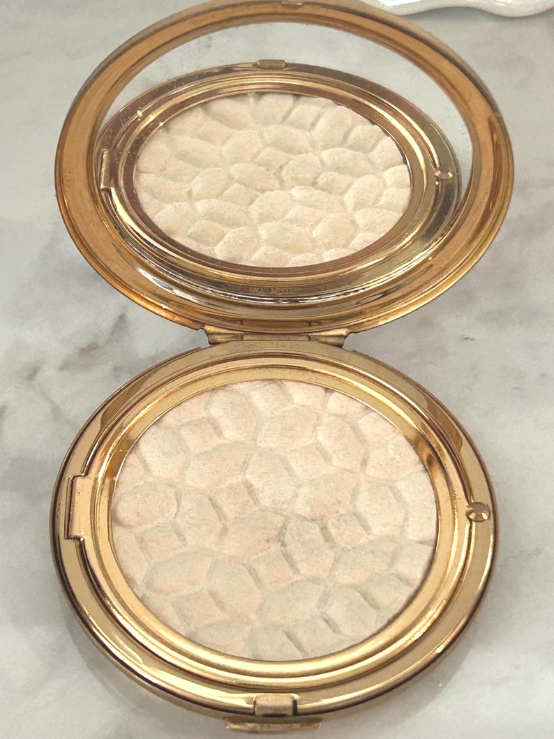 Peut inclure: Un bo&icirc;tier compact rond de couleur dor&eacute;e avec un couvercle &agrave; charni&egrave;re et un miroir int&eacute;gr&eacute;. L'int&eacute;rieur contient une poudre press&eacute;e pr&eacute;sentant un motif g&eacute;om&eacute;trique en relief rappelant un nid d'abeille, de couleur cr&egrave;me p&acirc;le.