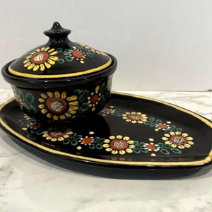 May include: A black ceramic lidded bowl and matching oval tray set. Both pieces feature a hand-painted floral pattern with yellow sunflowers, green leaves, and orange centers. The bowl has a rounded lid with a small knob handle and yellow trim.