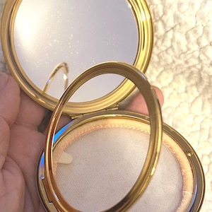 Peut inclure: Un poudrier rond en m&eacute;tal dor&eacute; avec un couvercle &agrave; charni&egrave;re. L'int&eacute;rieur pr&eacute;sente un miroir circulaire sur la moiti&eacute; sup&eacute;rieure et un tamis &agrave; poudre en maille blanche sur la moiti&eacute; inf&eacute;rieure. Un anneau m&eacute;tallique dor&eacute; repose sur le compartiment.