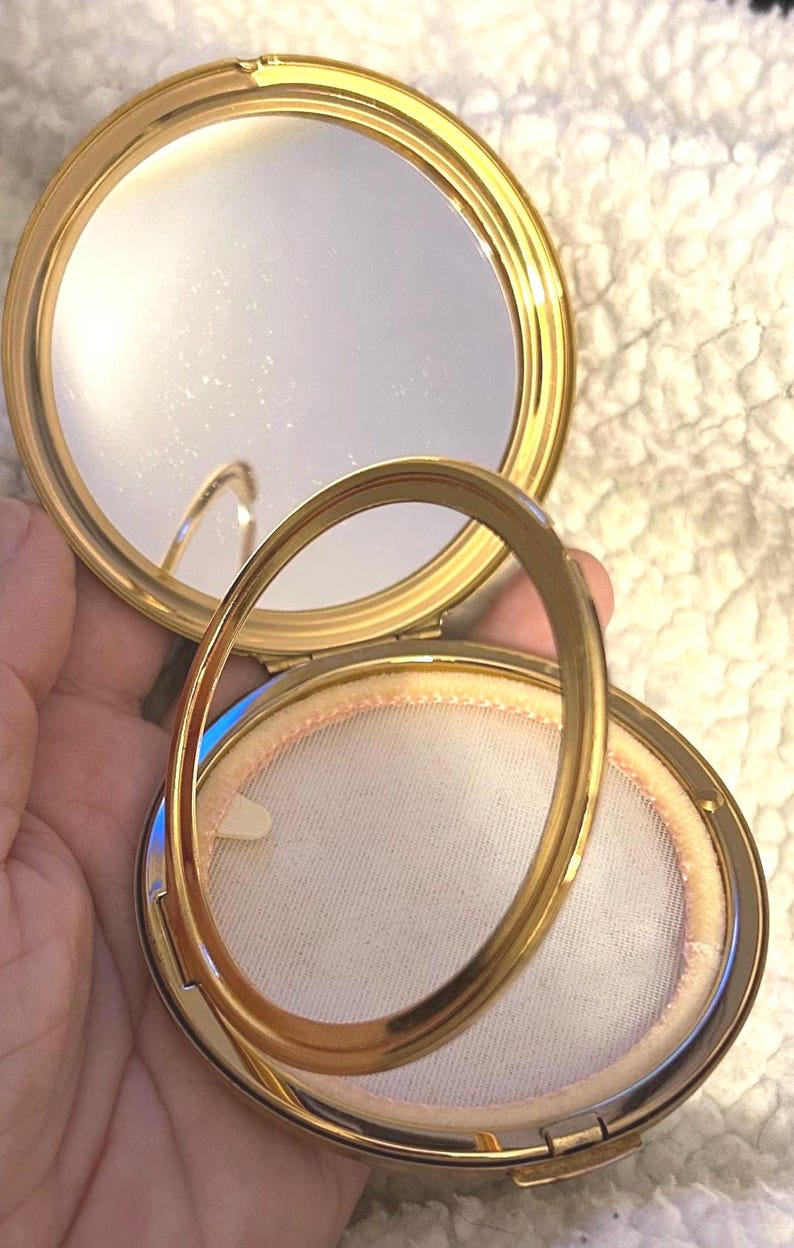 Peut inclure: Un poudrier rond en m&eacute;tal dor&eacute; avec un couvercle &agrave; charni&egrave;re. L'int&eacute;rieur pr&eacute;sente un miroir circulaire sur la moiti&eacute; sup&eacute;rieure et un tamis &agrave; poudre en maille blanche sur la moiti&eacute; inf&eacute;rieure. Un anneau m&eacute;tallique dor&eacute; repose sur le compartiment.