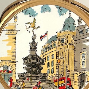 Peut inclure: Un poudrier rond dor&eacute; orn&eacute; d'une illustration color&eacute;e du monument de Piccadilly Circus &agrave; Londres, incluant la statue d'&Eacute;ros, un bus rouge &agrave; imp&eacute;riale et un drapeau Union Jack, avec le texte Piccadilly + Eros inscrit en bas.