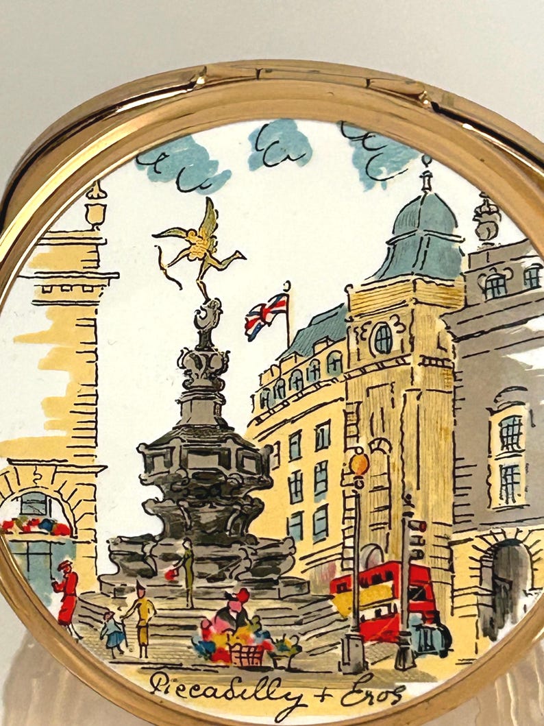 Peut inclure: Un poudrier rond dor&eacute; orn&eacute; d'une illustration color&eacute;e du monument de Piccadilly Circus &agrave; Londres, incluant la statue d'&Eacute;ros, un bus rouge &agrave; imp&eacute;riale et un drapeau Union Jack, avec le texte Piccadilly + Eros inscrit en bas.