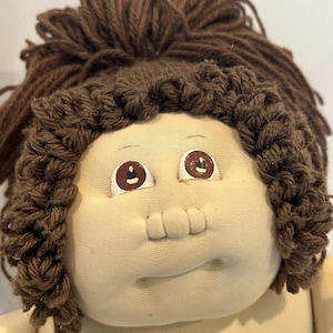 May include: Close-up of a vintage doll with a soft, fabric face and brown yarn hair. The doll has painted brown eyes and a small, stitched mouth. The doll's skin is a light beige color.