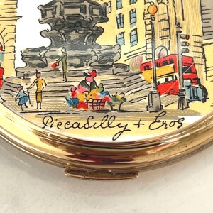 Peut inclure: Un miroir de poche rond dor&eacute; orn&eacute; d'une illustration color&eacute;e de la fontaine de Piccadilly Circus et de la statue d'Eros &agrave; Londres. La sc&egrave;ne montre un bus rouge &agrave; imp&eacute;riale et le texte manuscrit Piccadilly + Eros inscrit en bas.