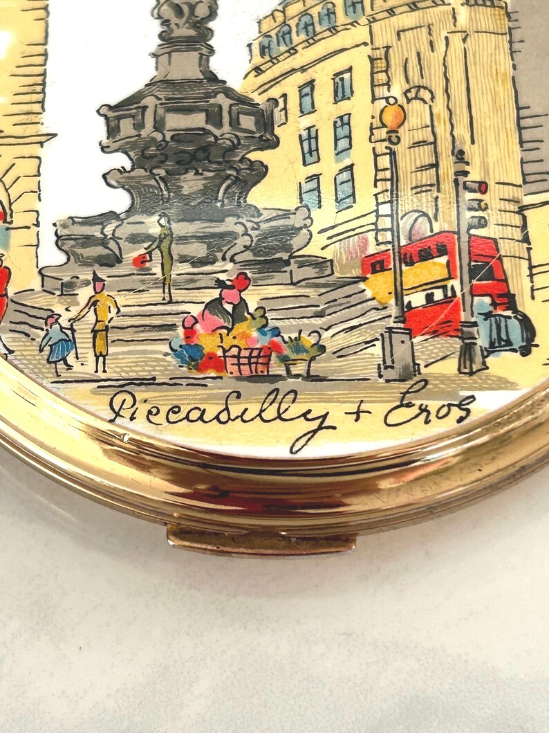 Peut inclure: Un miroir de poche rond dor&eacute; orn&eacute; d'une illustration color&eacute;e de la fontaine de Piccadilly Circus et de la statue d'Eros &agrave; Londres. La sc&egrave;ne montre un bus rouge &agrave; imp&eacute;riale et le texte manuscrit Piccadilly + Eros inscrit en bas.