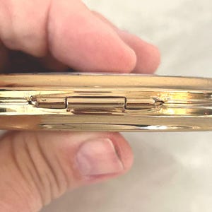 Peut inclure: Un bo&icirc;tier compact en m&eacute;tal dor&eacute; dot&eacute; d'un m&eacute;canisme de fermeture &agrave; charni&egrave;re. La surface ext&eacute;rieure est polie et r&eacute;fl&eacute;chissante, pr&eacute;sentant un profil de bord lisse et arrondi. Le design se compose de deux moiti&eacute;s circulaires reli&eacute;es par une charni&egrave;re centrale en m&eacute;tal.