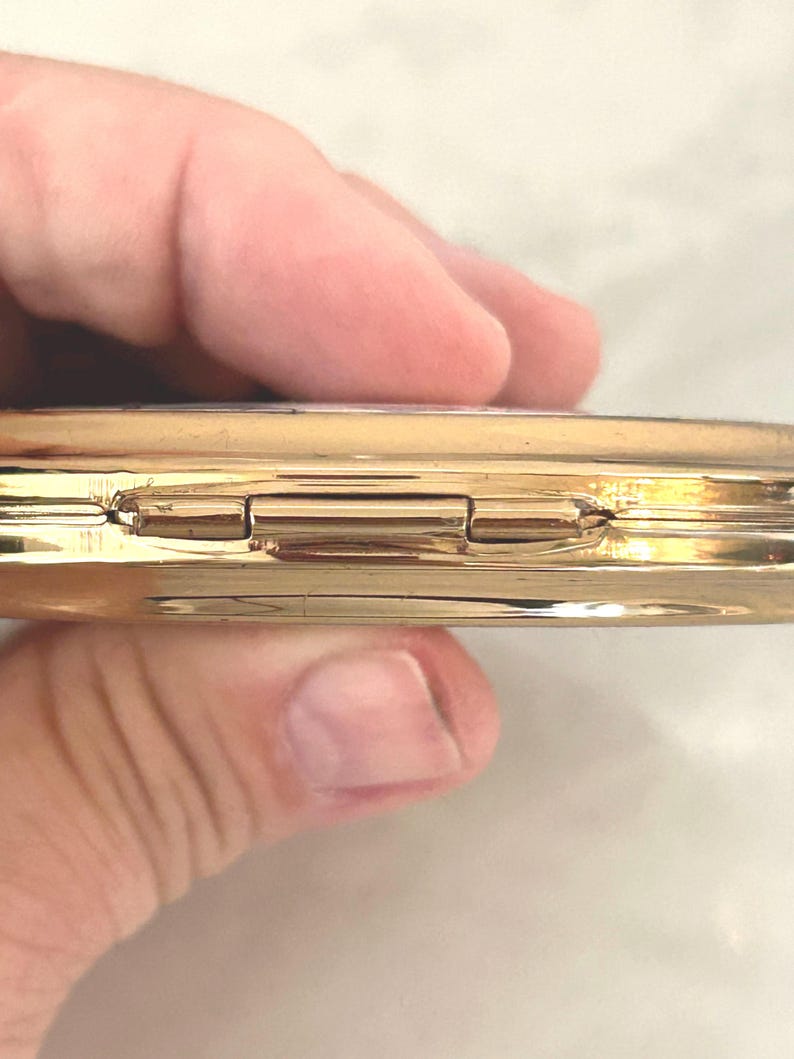 Peut inclure: Un bo&icirc;tier compact en m&eacute;tal dor&eacute; dot&eacute; d'un m&eacute;canisme de fermeture &agrave; charni&egrave;re. La surface ext&eacute;rieure est polie et r&eacute;fl&eacute;chissante, pr&eacute;sentant un profil de bord lisse et arrondi. Le design se compose de deux moiti&eacute;s circulaires reli&eacute;es par une charni&egrave;re centrale en m&eacute;tal.