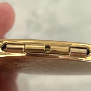 May include: A round, gold-toned metal compact mirror featuring a textured, patterned surface on the exterior. The object is shown from a side profile, highlighting the metallic hinge mechanism that connects the two halves of the circular case.