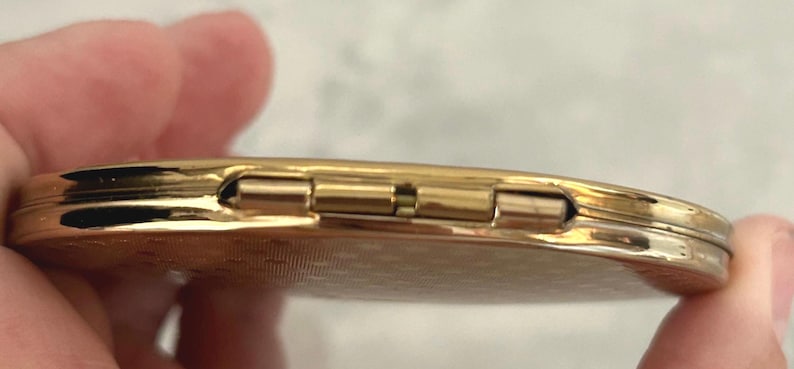 May include: A round, gold-toned metal compact mirror featuring a textured, patterned surface on the exterior. The object is shown from a side profile, highlighting the metallic hinge mechanism that connects the two halves of the circular case.