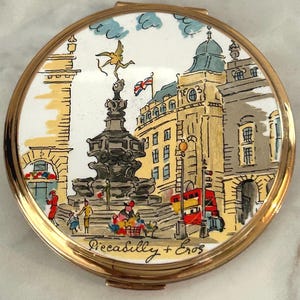 Peut inclure: Un poudrier rond en m&eacute;tal dor&eacute; orn&eacute; d'une illustration color&eacute;e du monument de Piccadilly Circus &agrave; Londres. La sc&egrave;ne repr&eacute;sente la statue d'&Eacute;ros, un bus rouge &agrave; imp&eacute;riale et le texte Piccadilly + Eros inscrit en bas de l'image.