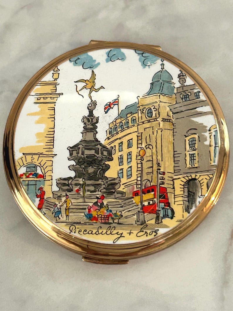 Peut inclure: Un poudrier rond en m&eacute;tal dor&eacute; orn&eacute; d'une illustration color&eacute;e du monument de Piccadilly Circus &agrave; Londres. La sc&egrave;ne repr&eacute;sente la statue d'&Eacute;ros, un bus rouge &agrave; imp&eacute;riale et le texte Piccadilly + Eros inscrit en bas de l'image.