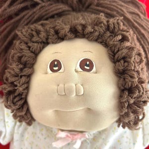 May include: A soft fabric doll with a beige face, brown yarn hair styled in a high ponytail, and brown painted eyes. The doll wears a white shirt featuring a small, multicolored floral pattern and a tiny pink ribbon at the center of the neckline.
