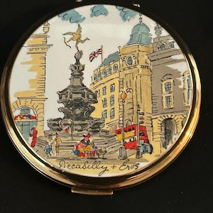 Peut inclure: Un miroir de poche rond en m&eacute;tal dor&eacute; orn&eacute; d'une illustration color&eacute;e de Piccadilly Circus &agrave; Londres. La sc&egrave;ne repr&eacute;sente la statue d'&Eacute;ros, un bus rouge &agrave; imp&eacute;riale et des b&acirc;timents, avec le texte manuscrit Piccadilly + Eros en bas.