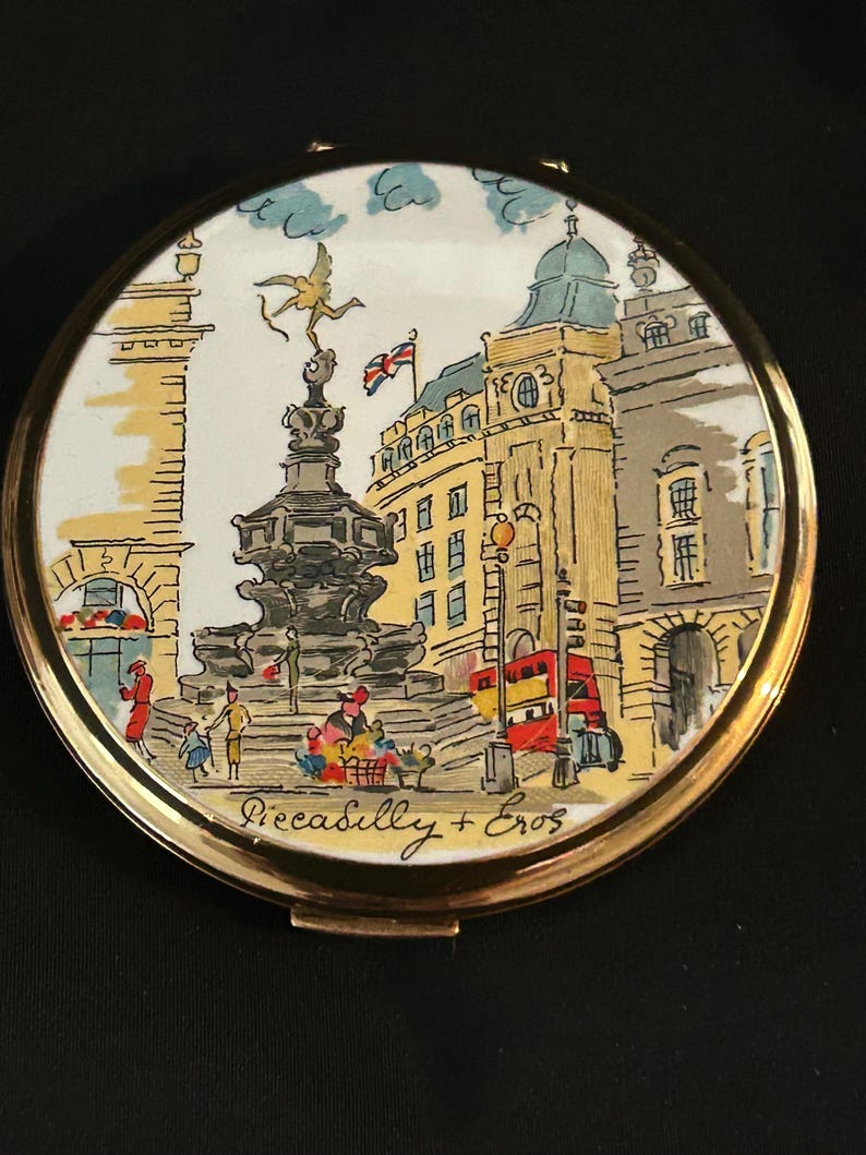 Peut inclure: Un miroir de poche rond en m&eacute;tal dor&eacute; orn&eacute; d'une illustration color&eacute;e de Piccadilly Circus &agrave; Londres. La sc&egrave;ne repr&eacute;sente la statue d'&Eacute;ros, un bus rouge &agrave; imp&eacute;riale et des b&acirc;timents, avec le texte manuscrit Piccadilly + Eros en bas.