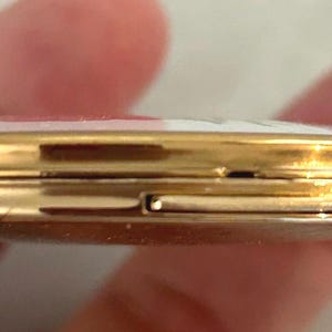 May include: A round, gold-toned metal compact case with a slim profile. The edges of the case are polished and reflective, showing a thin seam where the two halves meet. A small rectangular latch mechanism is visible on the side for opening.