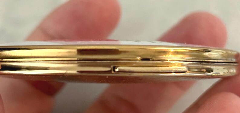May include: A round, gold-toned metal compact case with a slim profile. The edges of the case are polished and reflective, showing a thin seam where the two halves meet. A small rectangular latch mechanism is visible on the side for opening.