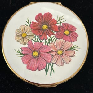 May include: A round gold-tone compact mirror featuring a white enamel lid decorated with a colorful illustration of five cosmos flowers in shades of red, pink, and cream with yellow centers and green stems against a plain white background.