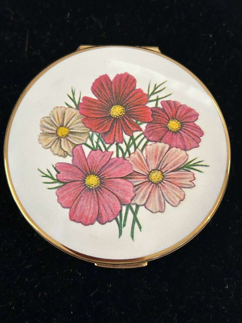 May include: A round gold-tone compact mirror featuring a white enamel lid decorated with a colorful illustration of five cosmos flowers in shades of red, pink, and cream with yellow centers and green stems against a plain white background.