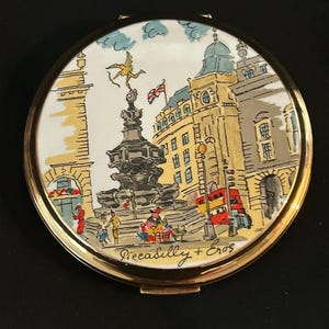Peut inclure: Un miroir de poche rond dor&eacute; orn&eacute; d'une illustration color&eacute;e du monument de Piccadilly Circus &agrave; Londres. La sc&egrave;ne repr&eacute;sente la statue d'&Eacute;ros, des b&acirc;timents environnants, un bus rouge &agrave; imp&eacute;riale et le texte Piccadilly + Eros.