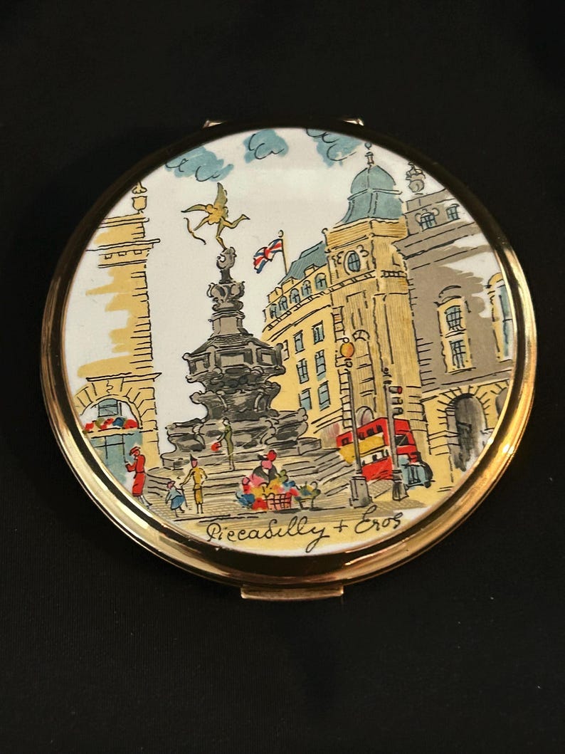 Peut inclure: Un miroir de poche rond dor&eacute; orn&eacute; d'une illustration color&eacute;e du monument de Piccadilly Circus &agrave; Londres. La sc&egrave;ne repr&eacute;sente la statue d'&Eacute;ros, des b&acirc;timents environnants, un bus rouge &agrave; imp&eacute;riale et le texte Piccadilly + Eros.