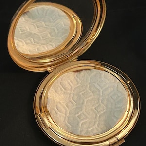 Peut inclure: Un poudrier rond en m&eacute;tal dor&eacute; avec un couvercle &agrave; charni&egrave;re et un miroir int&eacute;rieur. Le poudrier contient une poudre press&eacute;e avec un motif g&eacute;om&eacute;trique r&eacute;p&eacute;titif en relief sur toute sa surface, pr&eacute;sentant une finition textur&eacute;e blanc cass&eacute;.