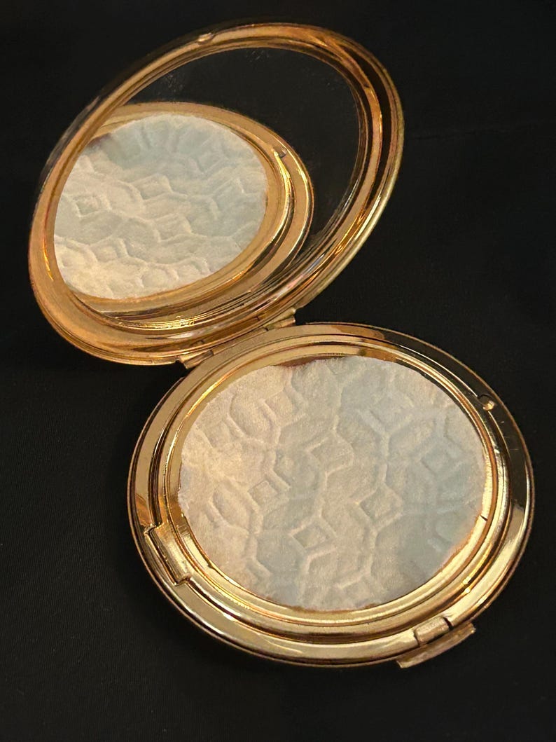 Peut inclure: Un poudrier rond en m&eacute;tal dor&eacute; avec un couvercle &agrave; charni&egrave;re et un miroir int&eacute;rieur. Le poudrier contient une poudre press&eacute;e avec un motif g&eacute;om&eacute;trique r&eacute;p&eacute;titif en relief sur toute sa surface, pr&eacute;sentant une finition textur&eacute;e blanc cass&eacute;.