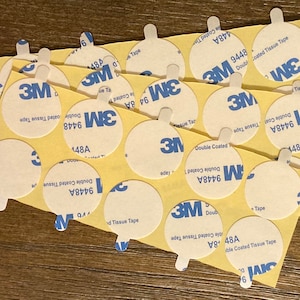 May include: A close-up of a pile of round, white, adhesive 3M double-coated tissue tape with yellow backing. The tape is labeled with the product number 9448A and 148A.
