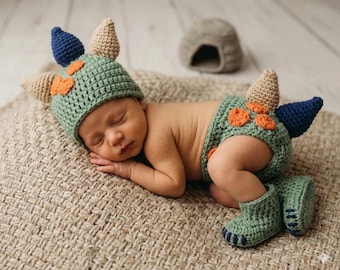 May include: A sleeping baby wearing a crocheted dinosaur costume. The costume includes a green hat with blue and tan spikes, matching diaper cover, and booties. Orange accents are on the hat and diaper cover. The baby is lying on a tan woven blanket.