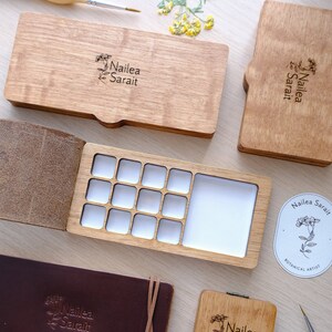 Puede incluir: Una paleta de acuarela de madera con 12 pocillos para pintura y un pocillo m&aacute;s grande para mezclar. La paleta est&aacute; abierta y tiene una cubierta de cuero. La paleta est&aacute; etiquetada como "Nailea Sarai Botanical Artist".