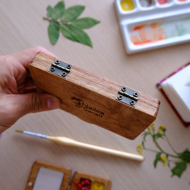 Puede incluir: Una caja de madera con tapa con bisagras, con dos bisagras de lat&oacute;n y el texto "Nailea Sarai" grabado en la parte delantera. La caja est&aacute; cerrada y se sostiene en una mano.