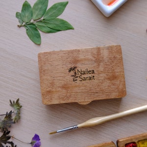 Puede incluir: Una caja de madera con tapa abatible, grabada con el texto "Nailea Sarai" y un dise&ntilde;o floral. Un pincel est&aacute; apoyado junto a la caja sobre una superficie de madera clara.
