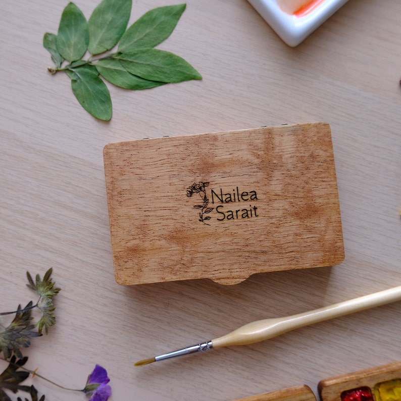 Puede incluir: Una caja de madera con tapa abatible, grabada con el texto "Nailea Sarai" y un dise&ntilde;o floral. Un pincel est&aacute; apoyado junto a la caja sobre una superficie de madera clara.