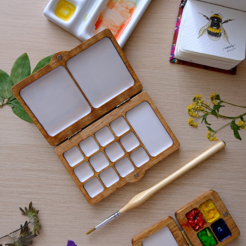 Puede incluir: Una paleta de acuarela de madera con 15 pocillos de pintura y dos &aacute;reas m&aacute;s grandes para mezclar pintura. La paleta est&aacute; abierta y tiene un pincel apoyado en ella. Hay una paleta de acuarela m&aacute;s peque&ntilde;a con cuatro pocillos en la esquina inferior derecha de la imagen.
