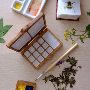Puede incluir: Una paleta de pintura de acuarela de madera con 14 pozos vac&iacute;os, abierta y lista para usar. La paleta est&aacute; hecha de madera marr&oacute;n claro y tiene un interior blanco. Un pincel est&aacute; acostado junto a la paleta.